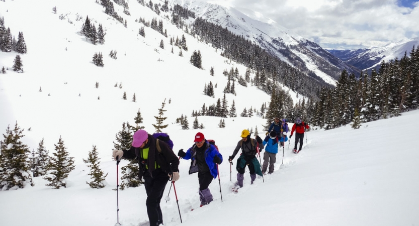 A group of people wearing winter gear hike in a line in a mountainous snowy landscape.